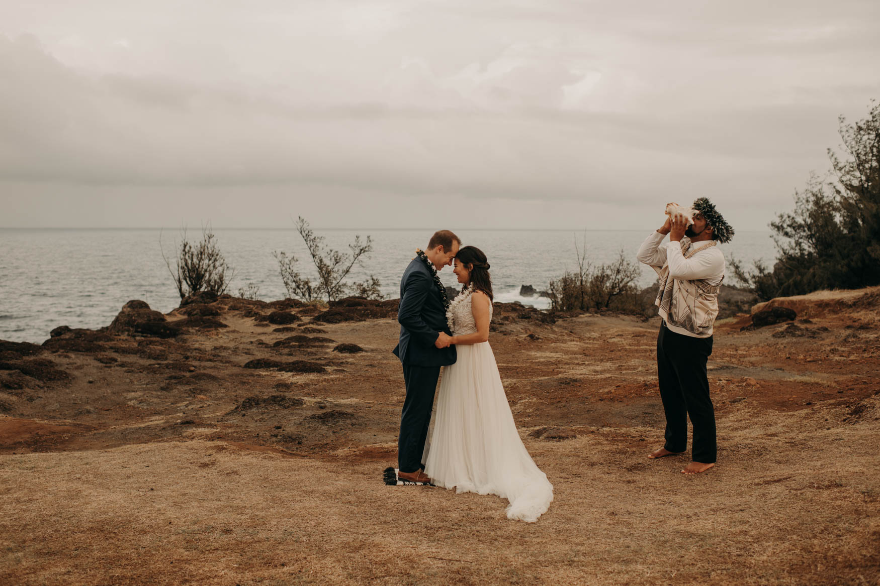 Andrew and Alison lean on one another, their forheads touching, while Euta blows the conch. The ocean and clouds behind them.