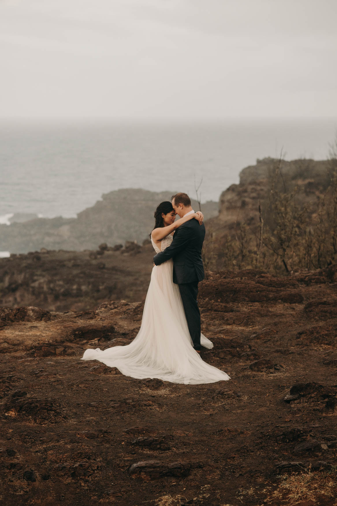 Andrew and Alison hug, Andrew's hands around her waist, her hands around his shoulders, their forheads touching. In the background are more cliffs, and further back the ocean.