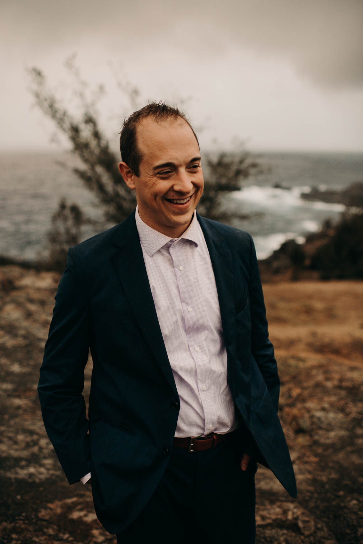 Andrew stands with his hands in the pockets of his dark blue suit, the buttons on the jacket all undone, and smiles while looking toward the right edge of the photo.