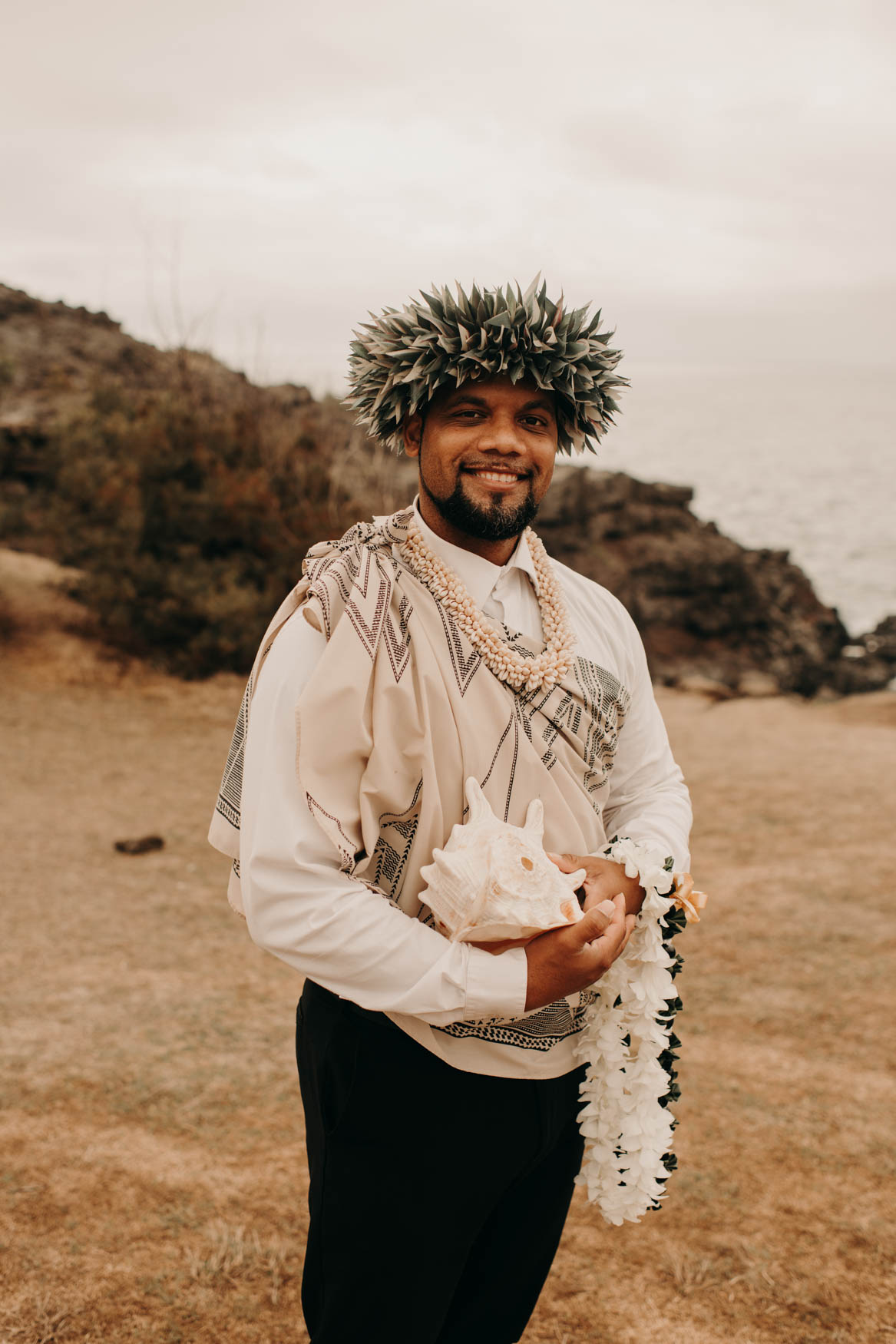 Euta, the officiant, smiles at the camera while holding a conch shell, and two leis 