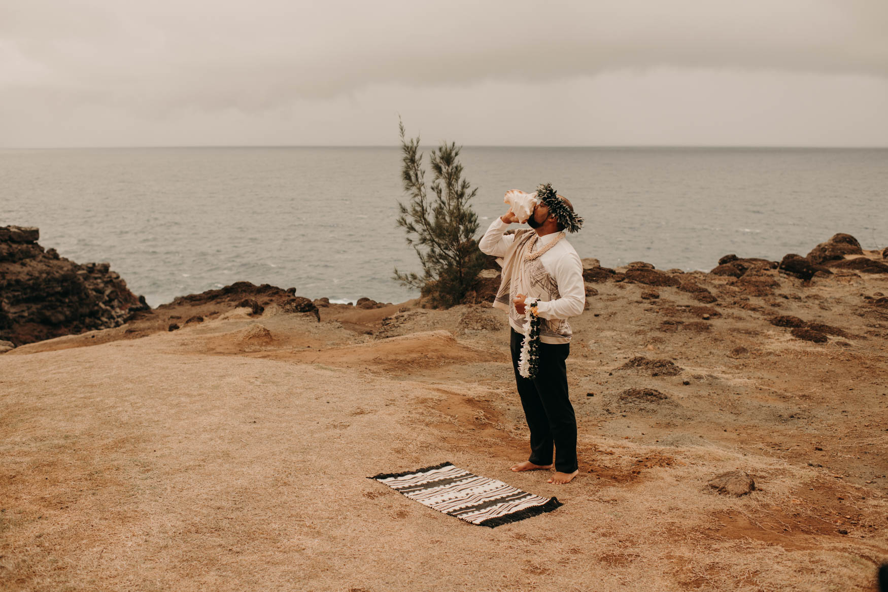 Euta stands with the ocean to his back, barefoot, and infront of a small rug, while blowing into the conch shell.